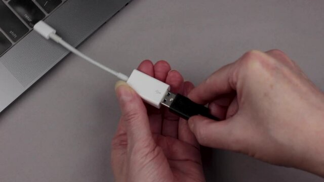 hands connecting a usb cable to a usb to usb-c adapter and then placing the adapter into a usb-c port of a computer, on a gray background