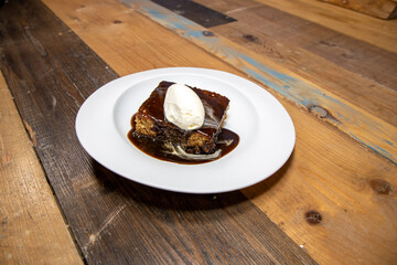 A white plate of delicious sticky toffee pudding chocolate cake with cream and chocolate sauce on a wooden kitchen work top