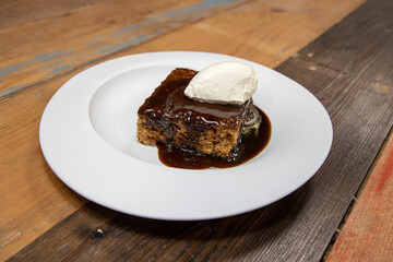 A white plate of delicious sticky toffee pudding chocolate cake with cream and chocolate sauce on a wooden kitchen work top