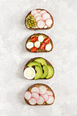 Vegetable sandwiches on slices of dark bread with radish, cucumber, cherry tomatoes, avocado, green peas flat lay on light concrete background