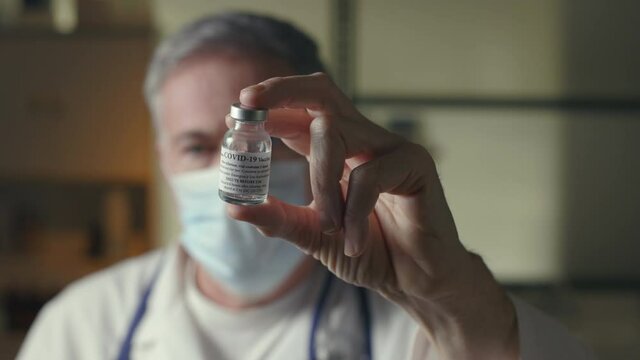 Scene rack focuses to a tiny vial of covid-19 vaccine from the doctor who displays it for the camera.