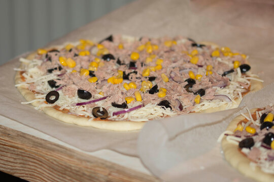 eine Pizza auf Backpapier frisch belegt mit Tunfisch, Maiskörnern, Oliven und weitern Zutaten 
