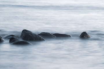 Fototapeta premium Long exposure of sea and rocks. Boulders sticking out from smooth wavy sea. Tranquil scene.