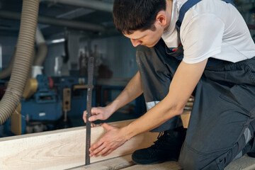 A young man works in a carpentry shop. An employee of Caucasian appearance in overalls measures wood with a ruler