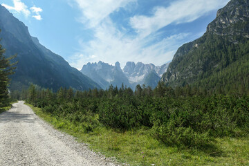 Fototapeta na wymiar A wide gravelled road in Italian Dolomites from the top of Strudelkopf leading towards the valley. A panoramic view on the high mountains. Sunny day. A few clouds above the peaks. Lush green plateau