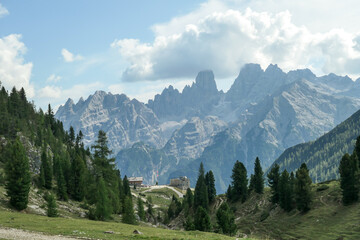 Fototapeta na wymiar A panoramic view on the high Italian Dolomites, hiding under the clouds. Sunny day. There is a small cottage at the end of a small pathway. Lush green plateau around. Few trees growing in between