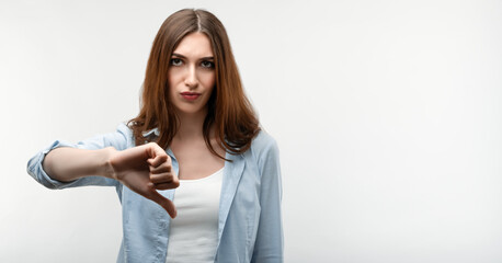 Beautiful caucasian girl with long chestnut hair, dressed in casual clothes, looking is upset at the camera, holding thumb down. Concept of dislike. Human emotions, facial expression concept