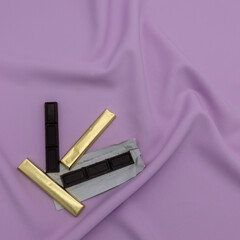 Four stick-shaped pieces of chocolate. Two are wrapped in golden paper. Lower left corner. Lilac background with folds.