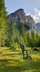 Fototapeta na wymiar A man with a big hiking backpack standing at the stump and enjoying the view on the high mountains around. There is a dense forest at the foothill and sharp, high mountains towering above. Calmness