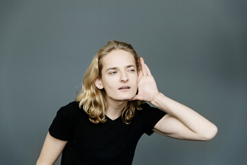 Attractive guy with long blonde hair, substituting a hand to his ear, eavesdrops with an interested...