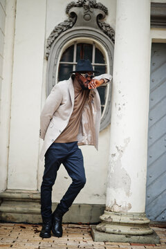 Stylish african american man wear beige jacket and  black hat with sunglasses pose against vintage window.