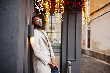 Stylish african american man wear beige jacket with sunglasses pose against house with golden garland.