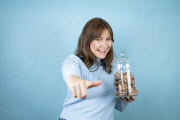 Young beautiful woman holding chocolate chips cookies jar over isolated blue background pointing to you and the camera with fingers, smiling positive and cheerful