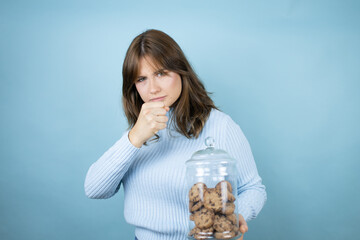 Young beautiful woman holding chocolate chips cookies jar over isolated blue background Punching fist to fight, aggressive and angry attack, threat and violence