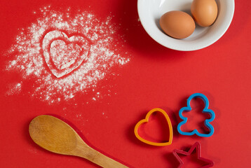 two heart-shaped prints scattered on a red background with white flour, next to plastic gingerbread molds. white plate with eggs and wooden spoon