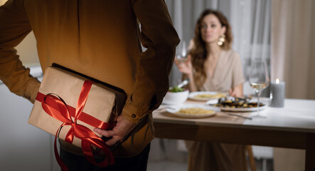 the woman is sitting at the table in a dress, the man is hiding the box behind his back. Gift concept for valentine's day, birthday,