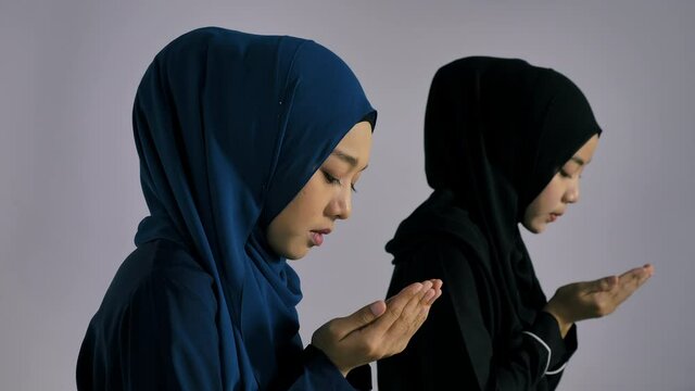 Two Asian Muslim young women in traditional hijab are praying glorify Allah and practicing the Islamic faith in mosque.