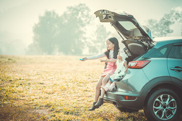 Girl and her infant sister sitting on back side of car playing with soap bubbles in park.