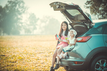 Naklejka premium Girl and her infant sister sitting on back side of car playing with soap bubbles in park.