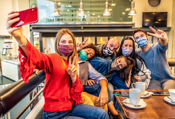 Young people having fun taking a selfie at coffee shop restaurant - New normal friendship concept with friends covered by face mask at restaurant