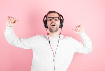 A funny man with a beard and a white sweatshirt listens to music with headphones, sings and dances on a pink background. A cheerful music lover listens to his favorite songs with headphones. Isolated.