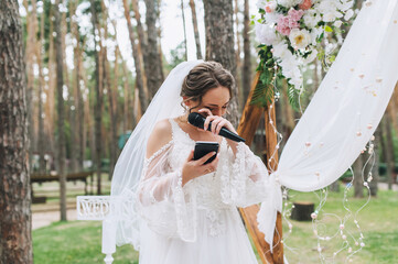 A bride in a white dress with a microphone and a smartphone in her hands is crying from the...