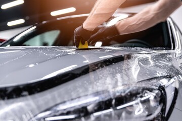 Car detailing studio worker applying protective ppf foil film on car body and lamps 