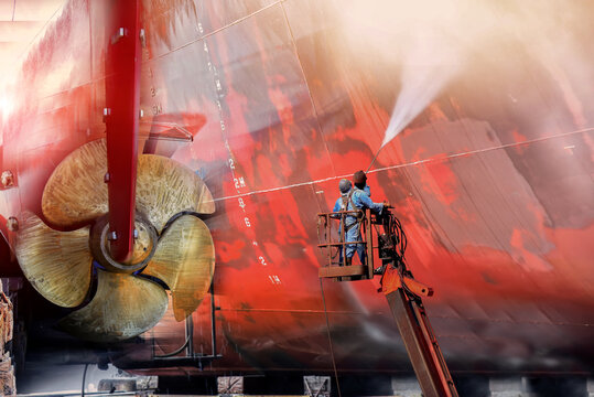 Worker in dry dock with water jet cleans the bottom of  the cargo ship from sea vegetation and muse colonies during routine overhaul in Shipyard and propeller background