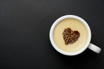 White mug of cappuccino, on a black background with copy space. A chocolate heart in the center of a cup of hot coffee drink.