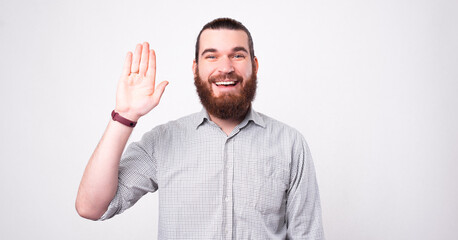 Fototapeta premium A happy young bearded man is waving and smiling at the same time at the camera near a white wall.