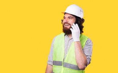 A photo of a cheerful young architect talking on his phone is looking away near a yellow wall .