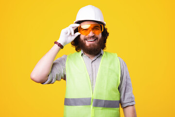 Young cheerful engineer is smiling at the camera and holding a pair of protective glasses is standing near a yellow wall .