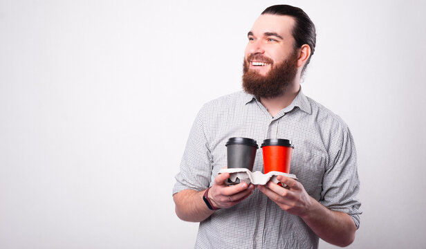 Cheerful young man is holding two hot drinks in papper cups smiling is looking away near a white wall .