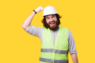 Excited bearded and young engineer is looking at the camera and is beating in the helmet thats on his head near a yellow wall .