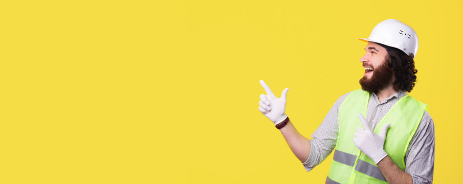 A bearded excited engineer is looking smiling and pointing at a free space near a yellow wall .