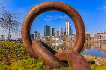 Naklejka premium Large metal ring with the skyline of Frankfurt. High-rise buildings of the city's financial district with river Main. Park along the shore in sunny weather with blue sky and reflections in the water