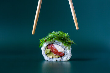 Chopsticks are going to take roll with tuna and chuka seaweed on a dark green background