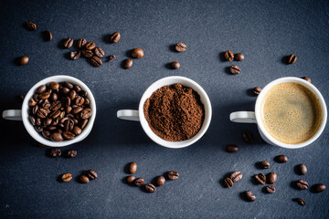 3 Tassen mit Kaffeebohnen, Kaffeepulver, Kaffee und einzelne Bohnen auf einer Schiefertafel