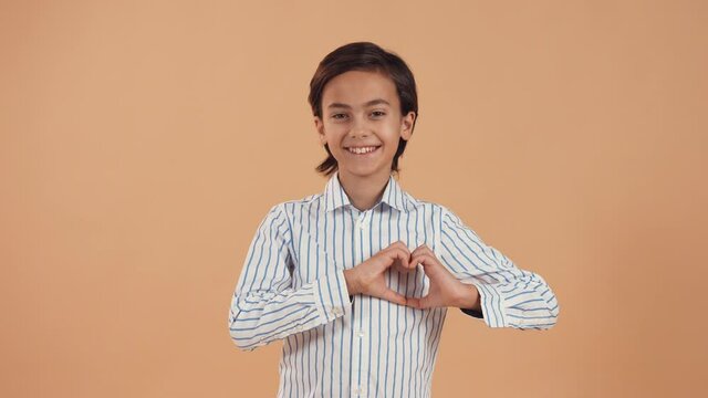 Cute smiling caucasian boy in a white striped shirt shows a heart-shaped gesture with his hands isolated on a peach tender background. Valentine's Day.