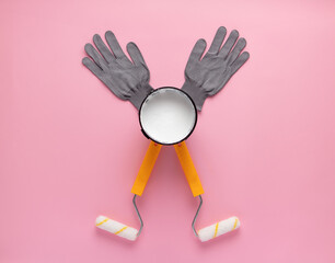 Fototapeta premium Creative composition of paint rollers, open can of white paint, and work gloves on a pink background. Vizualization of happy man with raised hands doing house repairs.