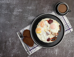 Fried eggs with pepper and sausage and a cup of coffee on on a round plate on a dark background. Top view, flat lay