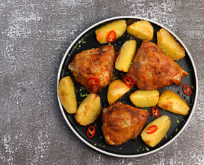 Chicken thigh with potatoes and hot peppers on a round plate on a dark background. Top view, flat lay