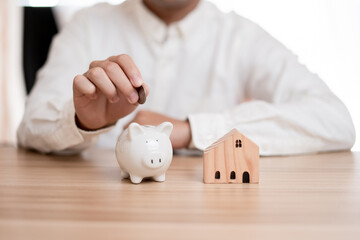  Hand of businessman putting coin in Little Piggy Bank and small wooden toy house placed on the...