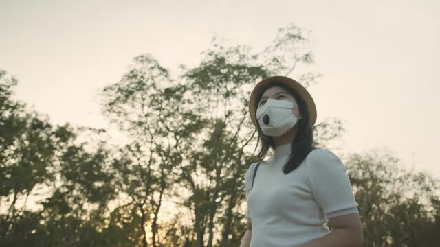 social distancing safety travel new normal lifestyle asian female woman wearing protective face mask walking and take photo with smartphone tour in public garden park sunset moment