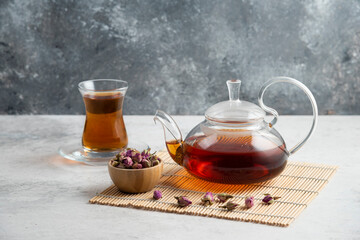 A glass cup of tea with dried roses and teapot