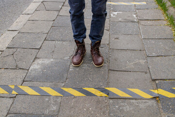 Male feet in boots on the sidewalk in front of a black and yellow stripe..