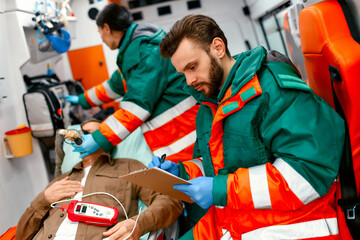 Fototapeta premium A paramedic woman in uniform puts on a ventilator with oxygen to help senior patient lying with a pulse oximeter on a gurney in modern ambulance. The male paramedic takes notes on the patient's card.