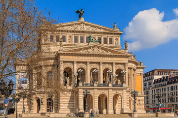 Obraz premium Buildings of the historic old opera house with trees and commercial buildings at springtime in sunshine with clouds in the sky. Square in the center of the city of Frankfurt.
