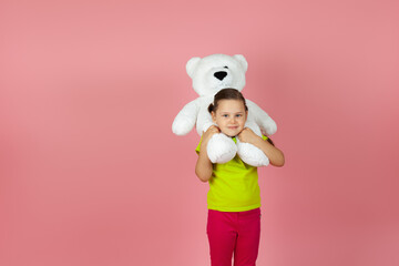 a beautiful child with pigtails in a light green T-shirt put a white teddy bear on her shoulders , isolated on a pink background.