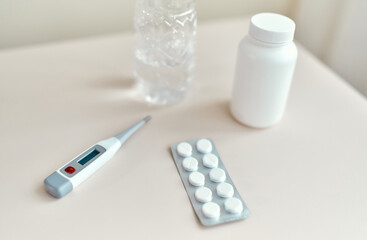 Close-up image of a thermometer, pills, a jar of medicine and a glass of water lying on the table.